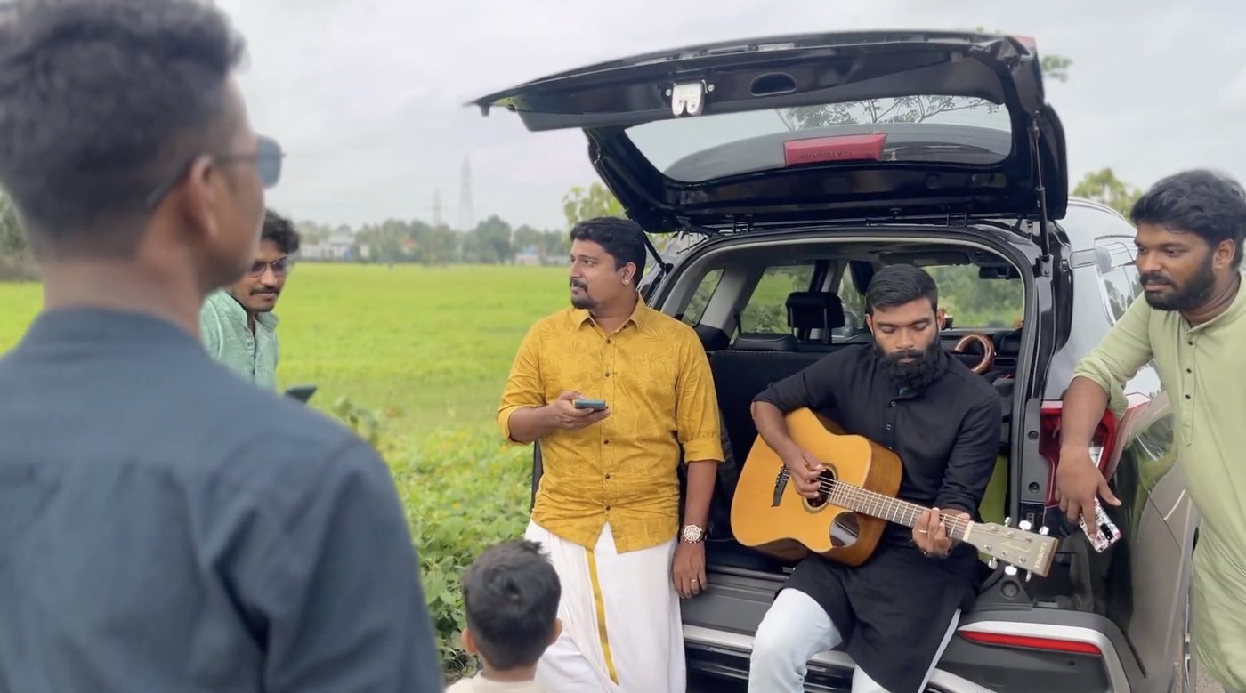 The team hanging out by an open car boot in a green field, one of them playing the guitar